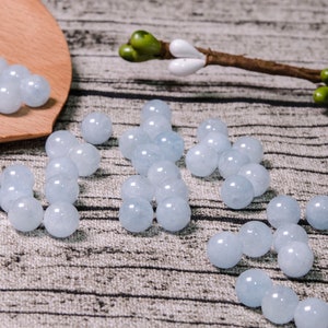 May include: A collection of light blue round beads, some are arranged in a line, others are in a pile on a wooden spoon. The beads are on a textured surface.
