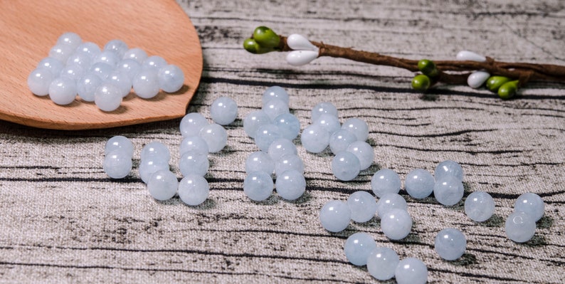 May include: A collection of light blue round beads, some are arranged in a line, others are in a pile on a wooden spoon. The beads are on a textured surface.