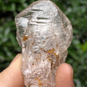 Puede incluir: Un cristal de cuarzo en bruto y transparente con una superficie rugosa y texturizada. El cristal tiene inclusiones de minerales de color marrón dorado y se sostiene en una mano sobre un fondo verde borroso. El cuarzo tiene forma irregular y refleja la luz.