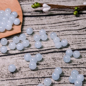 May include: A collection of light blue round beads, some are in a wooden spoon, and others are scattered on a textured surface.