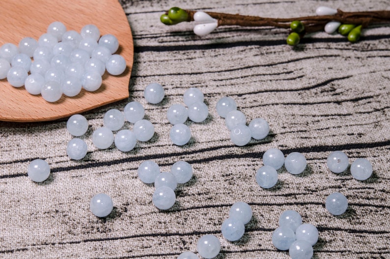 May include: A collection of light blue round beads, some are in a wooden spoon, and others are scattered on a textured surface.