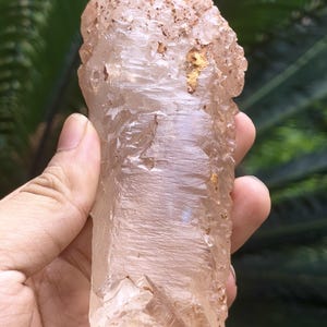 Può includere: Un esemplare di cristallo di quarzo rosa pallido traslucido con una struttura allungata e frastagliata. La superficie presenta striature naturali, sfaccettature cristalline e piccole macchie di depositi minerali terrosi bruno-aranciati.