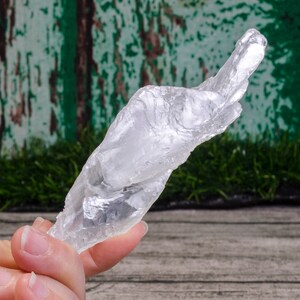 Può includere: Una punta di cristallo trasparente e bianca con una forma unica e contorta. Il cristallo è tenuto in una mano su uno sfondo verde e marrone.