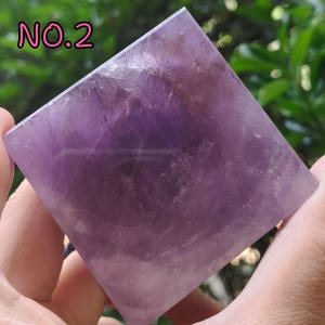May include: A square-shaped amethyst crystal pyramid. The pyramid is a deep purple color with lighter streaks and variations in tone. The text "NO.2" is in pink letters in the upper left corner. The pyramid is held in a person's hand.