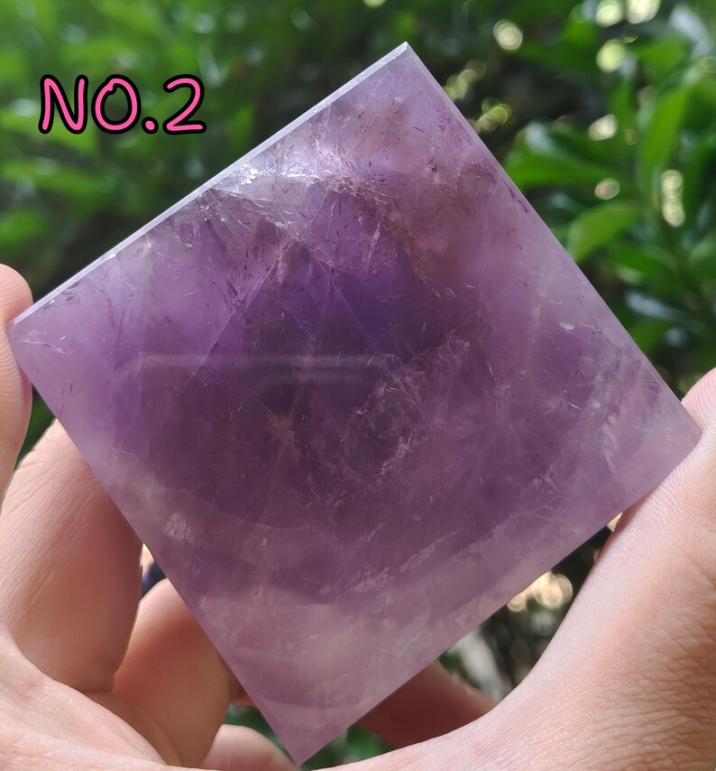 May include: A square-shaped amethyst crystal pyramid. The pyramid is a deep purple color with lighter streaks and variations in tone. The text "NO.2" is in pink letters in the upper left corner. The pyramid is held in a person's hand.