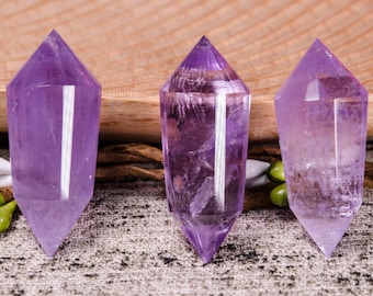 May include: Three amethyst crystal points, each with a pointed tip and a clear, translucent purple color. The crystals are arranged in a row on a textured surface.