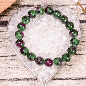 May include: A green and pink gemstone bracelet on a heart-shaped background. The bracelet is made of round beads and is lying on a bed of white stones.