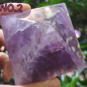 May include: A pyramid-shaped amethyst crystal, displaying shades of purple and translucent areas. The crystal is held in a hand, with the text "NO.2" in pink at the top left. The pyramid's facets are visible, showcasing the natural formation and texture of the stone.
