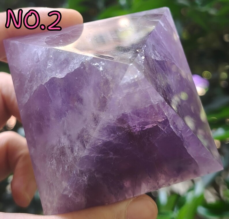 May include: A pyramid-shaped amethyst crystal, displaying shades of purple and translucent areas. The crystal is held in a hand, with the text "NO.2" in pink at the top left. The pyramid's facets are visible, showcasing the natural formation and texture of the stone.