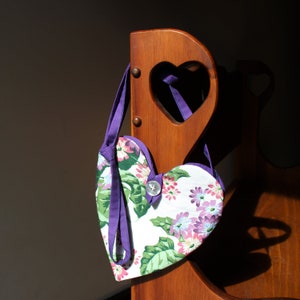 May include: A heart-shaped fabric bag with a floral print hangs on a wooden heart-shaped cutout. The bag has a purple strap and a white button.