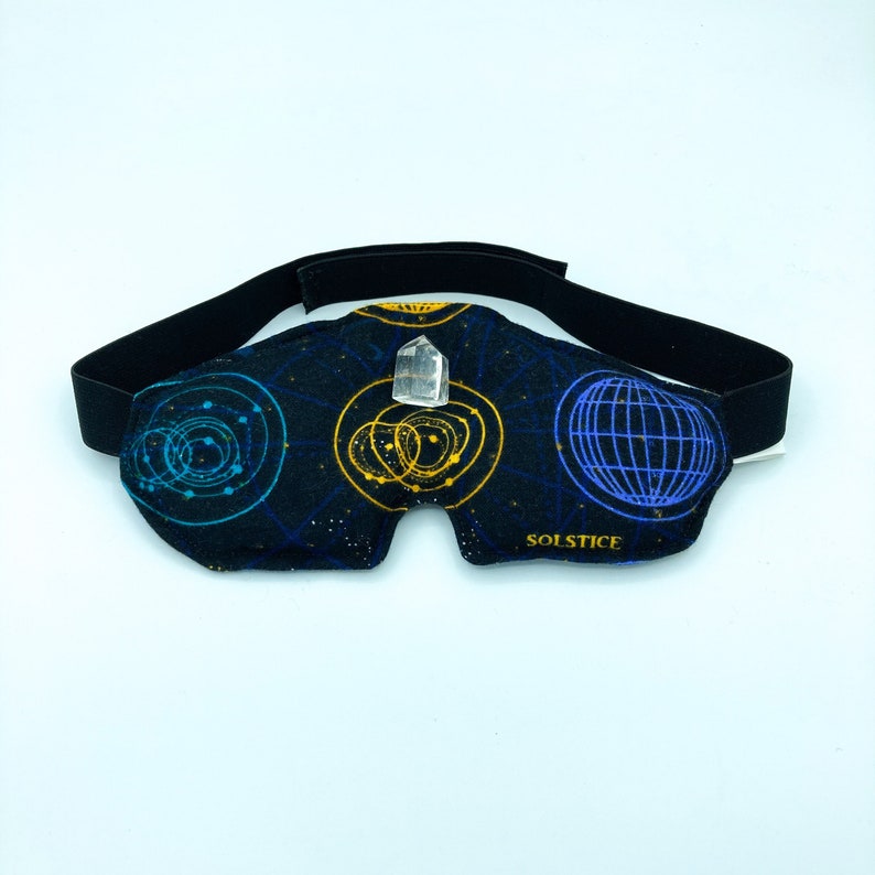 May include: A black eye mask with a black elastic strap. The mask features a celestial design in blue and yellow, with the word "SOLSTICE" printed in gold. A clear crystal is attached to the center of the mask.