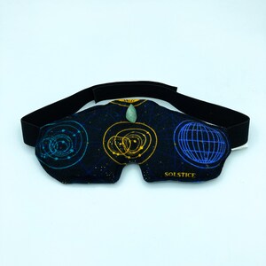 May include: Black eye mask with a celestial design featuring blue and gold geometric patterns. The mask has a black elastic strap and a small green stone at the center. The word "SOLSTICE" is printed on the mask.