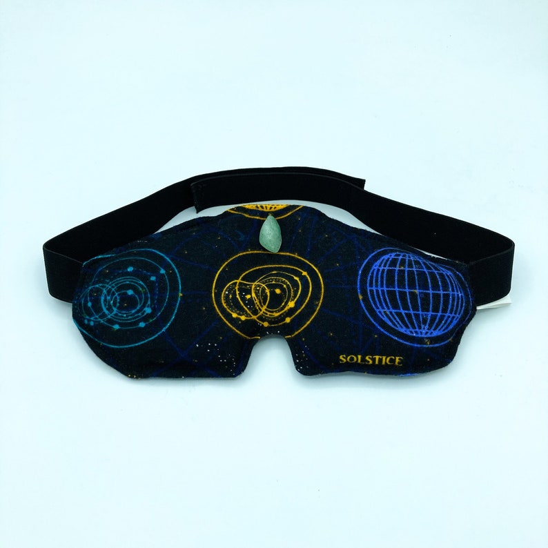 May include: Black eye mask with a celestial design featuring blue and gold geometric patterns. The mask has a black elastic strap and a small green stone at the center. The word "SOLSTICE" is printed on the mask.