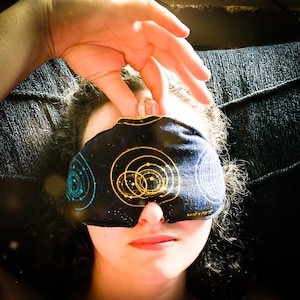 May include: A black sleep mask with a celestial design featuring gold and teal planets and stars. A clear crystal is held above the mask. The mask is covering the eyes.