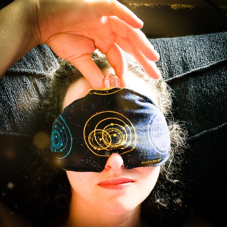 May include: A black sleep mask with a celestial design featuring gold and teal planets and stars. A clear crystal is held above the mask. The mask is covering the eyes.