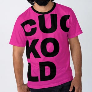 Op de afbeelding: Een felroze t-shirt met het woord "CUCKOLD" in grote, zwarte letters. Het shirt heeft een zwarte kraag en korte mouwen. De tekst is verticaal uitgelijnd en beslaat het grootste deel van de voorkant van het shirt.