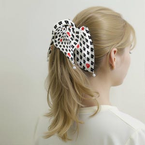 May include: A white hair scarf with a playing card design, featuring black spades and red hearts. The scarf is tied in a bow and adorned with small, white pearl accents. The hair accessory is set against a background of blonde hair.