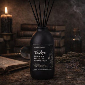 May include: A black essential oil reed diffuser with black reeds. The label reads "Thicket" with the scents of Lavender, Rosemary, and Grapefruit. The bottle is 200ml. The background includes books and a lit candle.