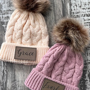 May include: Two knitted cable knit beanies with faux fur pom poms. The beanies are in cream and pink with brown leather patches that say "Grace" and "Everly".