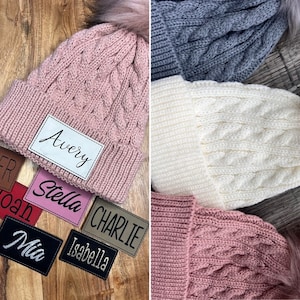 May include: A collection of knitted hats with personalized name tags. The hats are in various colors, including pink, white, and gray. The name tags are made of leather and have names such as Avery, Stella, Charlie, Isabella, and Mia.