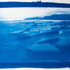 May include: A cyanotype print of a coastal landscape. The image features a shoreline with large, angular rocks in the foreground, a calm sea, and a distant coastline under a bright sky. The print is framed with a dark blue border.