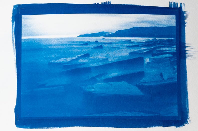 May include: A cyanotype print of a coastal landscape. The image features a shoreline with large, angular rocks in the foreground, a calm sea, and a distant coastline under a bright sky. The print is framed with a dark blue border.