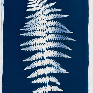 May include: A cyanotype print of a fern frond against a deep blue background. The fern is rendered in white, showcasing intricate details of the leaves. The print is on a white paper with text at the bottom.