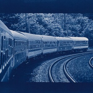 May include: A cyanotype print of a train traveling along curved tracks. The train cars are detailed with multiple windows, and the scene is set against a backdrop of trees. The image has a vintage, artistic feel.