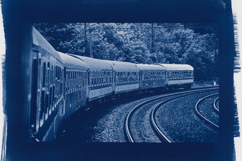 May include: A cyanotype print of a train traveling along curved tracks. The train cars are detailed with multiple windows, and the scene is set against a backdrop of trees. The image has a vintage, artistic feel.
