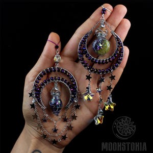 May include: A pair of crescent moon earrings with dangling stars and a small glass vial filled with iridescent glitter. The earrings are made with silver wire and purple beads.