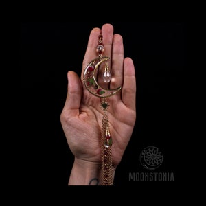 May include: A gold crescent moon charm with a clear crystal drop and a red and green floral design inside. The charm is attached to a gold chain with a tassel.