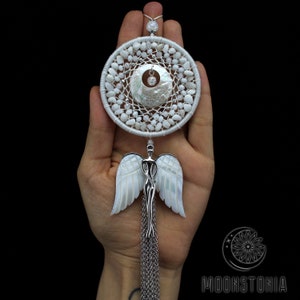 May include: A white dreamcatcher with a central circular design, featuring a web of white thread, small white stones, and a mother-of-pearl accent. Below, a silver angel pendant with mother-of-pearl wings hangs from a chain.