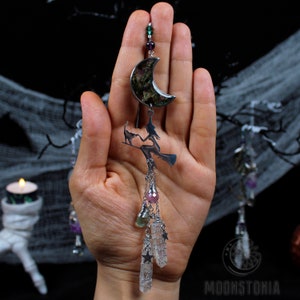 May include: A silver charm necklace with a crescent moon pendant featuring a green and purple resin design. The charm hangs from a chain with a witch and cat silhouette, and a cluster of clear quartz crystals with silver stars.