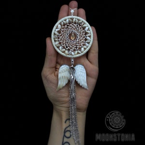 May include: White dreamcatcher with a silver angel charm and a beaded fringe. The dreamcatcher is decorated with white and brown seashells.