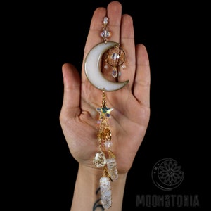 May include: A white crescent moon charm with a gold-tone chain and crystal accents. The charm features a small dreamcatcher design and a star-shaped pendant. The chain is adorned with crystals and beads.