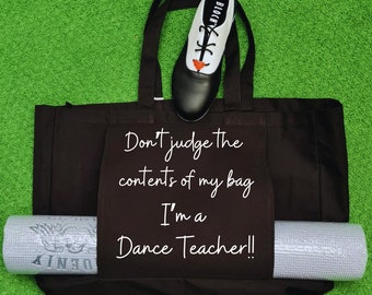 May include: A black tote bag with the text "Don't judge the contents of my bag I'm a Dance Teacher!!" printed on it. A white and black dance shoe is on top of the bag, and a gray yoga mat is sticking out of the side pocket.