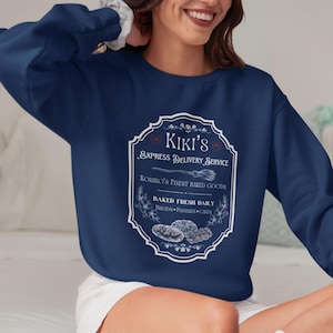Puede incluir: Sudadera azul marino con un gr&aacute;fico blanco y azul. El gr&aacute;fico dice "Kiki's Express Delivery Service" con texto sobre productos horneados. La sudadera tiene cuello redondo y mangas largas.