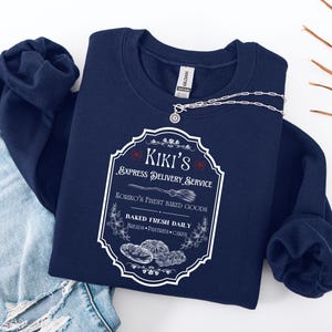 Puede incluir: Una sudadera azul marino con un gr&aacute;fico blanco que presenta el texto "Kiki's Express Delivery Service" y "Koriko's Finest Baked Goods". El dise&ntilde;o incluye ilustraciones de productos horneados y una escoba. Un collar plateado est&aacute; sobre la sudadera.