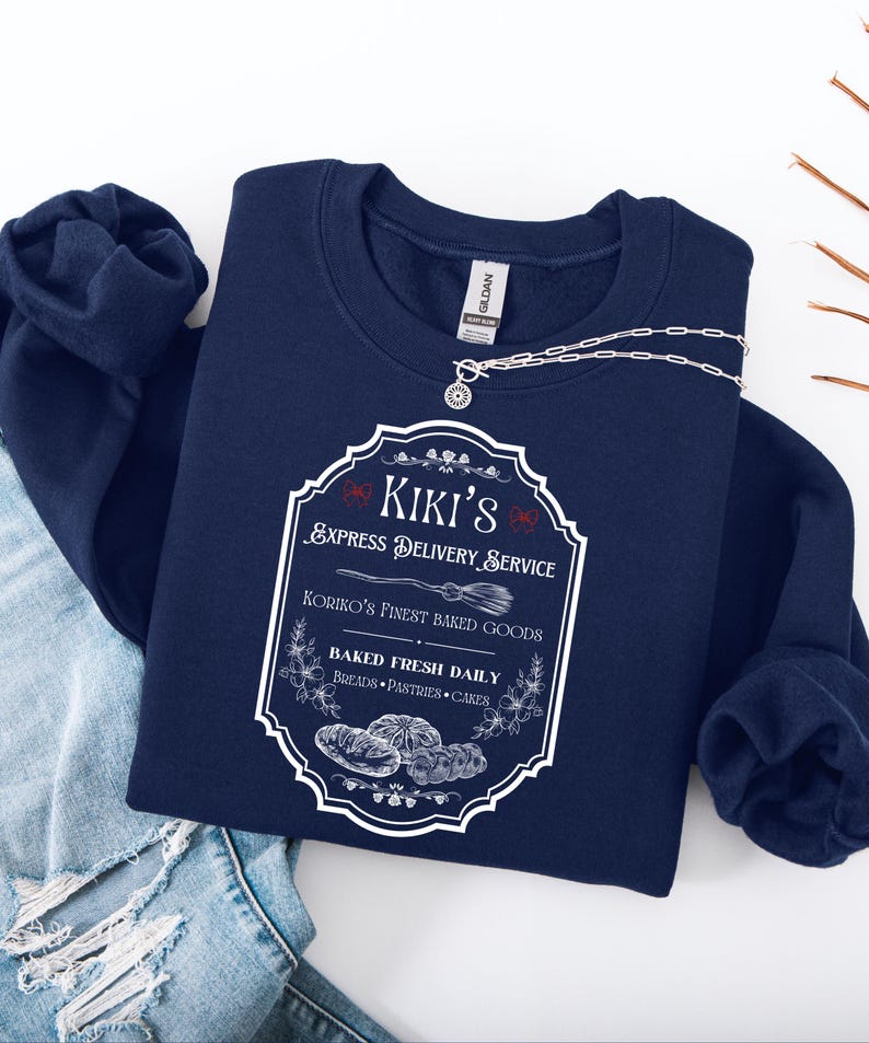 Puede incluir: Una sudadera azul marino con un gr&aacute;fico blanco que presenta el texto "Kiki's Express Delivery Service" y "Koriko's Finest Baked Goods". El dise&ntilde;o incluye ilustraciones de productos horneados y una escoba. Un collar plateado est&aacute; sobre la sudadera.