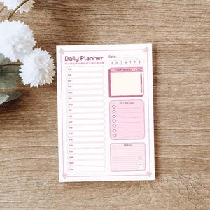 May include: A pink and white daily planner with a pink and white computer icon, a to-do list, and a notes section. The planner has a pink flower design and the text "Daily Planner" at the top.