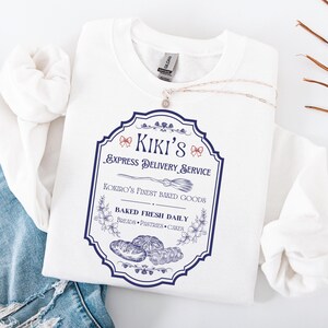 Puede incluir: Una sudadera blanca con un gr&aacute;fico azul y blanco que dice "Kiki's Express Delivery Service" con una ilustraci&oacute;n de una escoba. El gr&aacute;fico tambi&eacute;n dice "Kokiro's Finest Baked Goods" y "Baked Fresh Daily". Un collar plateado est&aacute; sobre la sudadera.