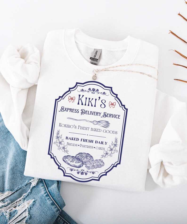 Puede incluir: Una sudadera blanca con un gr&aacute;fico azul y blanco que dice "Kiki's Express Delivery Service" con una ilustraci&oacute;n de una escoba. El gr&aacute;fico tambi&eacute;n dice "Kokiro's Finest Baked Goods" y "Baked Fresh Daily". Un collar plateado est&aacute; sobre la sudadera.