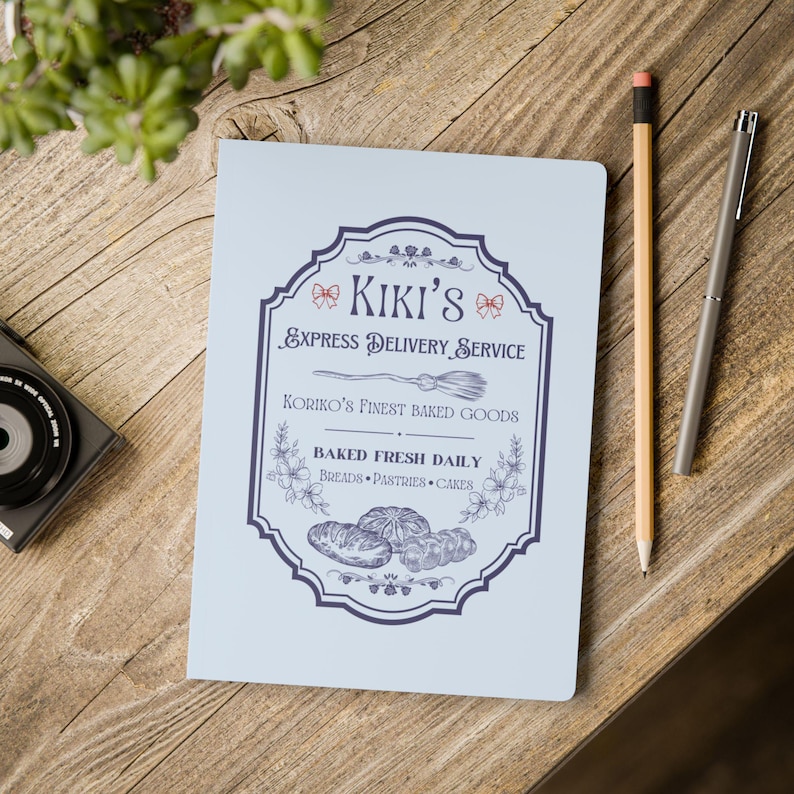 May include: A light blue notebook with a vintage-style logo that says "Kiki's Express Delivery Service" and "Koriiko's Finest Baked Goods". The logo also includes a broom, a bow, and a floral design. The text "Baked Fresh Daily" is also included in the logo.