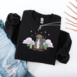 Puede incluir: Sudadera negra con una ilustración de dibujos animados de una ardilla con un sombrero de vaquero y botas. La ardilla está de pie frente a un paisaje desértico con cactus y estrellas.