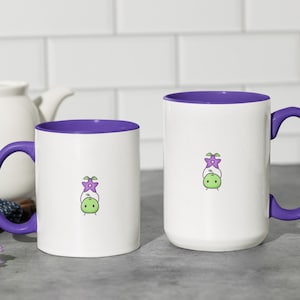 Könnte beinhalten: Zwei weiße Keramikbecher mit violetten Rändern und Griffen. Beide Becher zeigen eine Cartoon-Illustration eines grünen Wesens mit einer violetten Blume auf dem Kopf. Die Becher stehen auf einer grauen Oberfläche.
