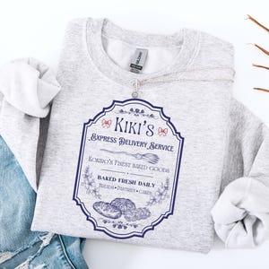 Puede incluir: Una sudadera gris claro con un gr&aacute;fico azul y blanco que dice "Kiki's Express Delivery Service". El gr&aacute;fico incluye una escoba, flores e ilustraciones de productos horneados. La sudadera est&aacute; doblada sobre un par de vaqueros azules.