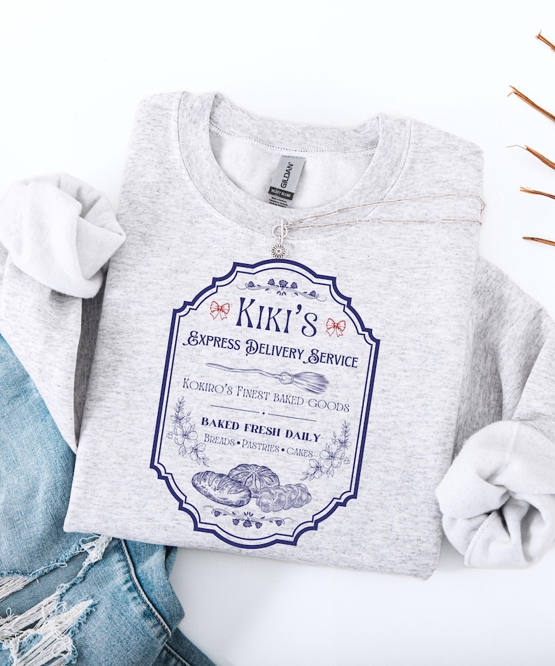 Puede incluir: Una sudadera gris claro con un gr&aacute;fico azul y blanco que dice "Kiki's Express Delivery Service". El gr&aacute;fico incluye una escoba, flores e ilustraciones de productos horneados. La sudadera est&aacute; doblada sobre un par de vaqueros azules.