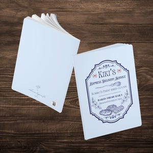 May include: Two light blue notebooks with a vintage design. The front cover of one notebook features a logo with the text "Kiki's Express Delivery Service" and the text "Koriro's Finest Baked Goods Baked Fresh Daily Breads, Pastries, Cakes".