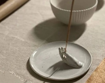 May include: White ceramic incense holder shaped like a hand with a white incense stick burning in the palm of the hand. The holder is on a white ceramic plate.