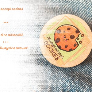 Op de afbeelding: Een ronde houten pin met een cartoon-cookie-ontwerp. De cookie heeft chocolade chips en een glimlachend gezicht. De tekst "Always accept COOKIES" staat rond de cookie. De pin is op een blauwe denim achtergrond.